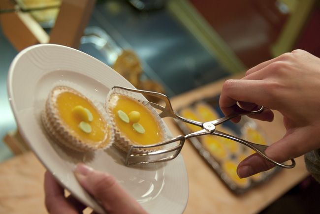 Entreprise - Photographie en plan rapproché illustrant la gestuelle de servir une tartelette au citron avec une pince en métal pour la boulangerie confiserie Le Colibri